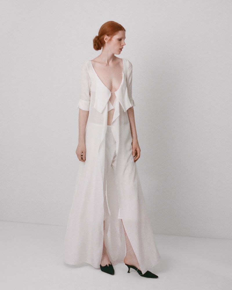 Model in long white sheer coat and trousers standing against a plain white studio backdrop.