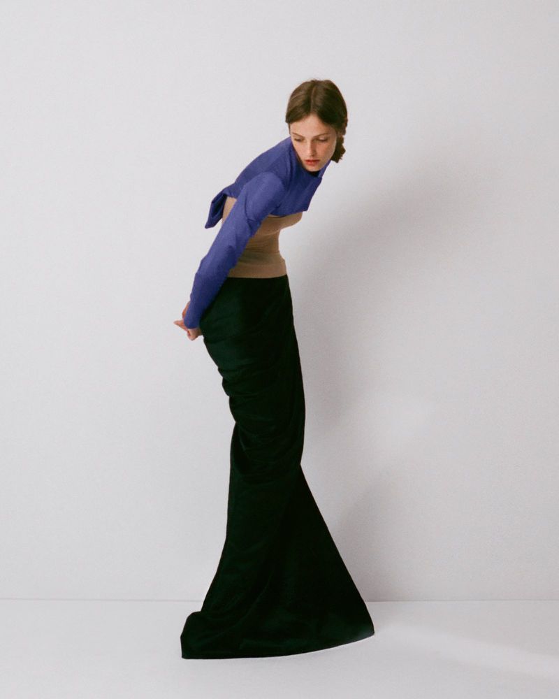 Model wearing a blue top, beige corset, and long black skirt against a plain white seamless studio background.