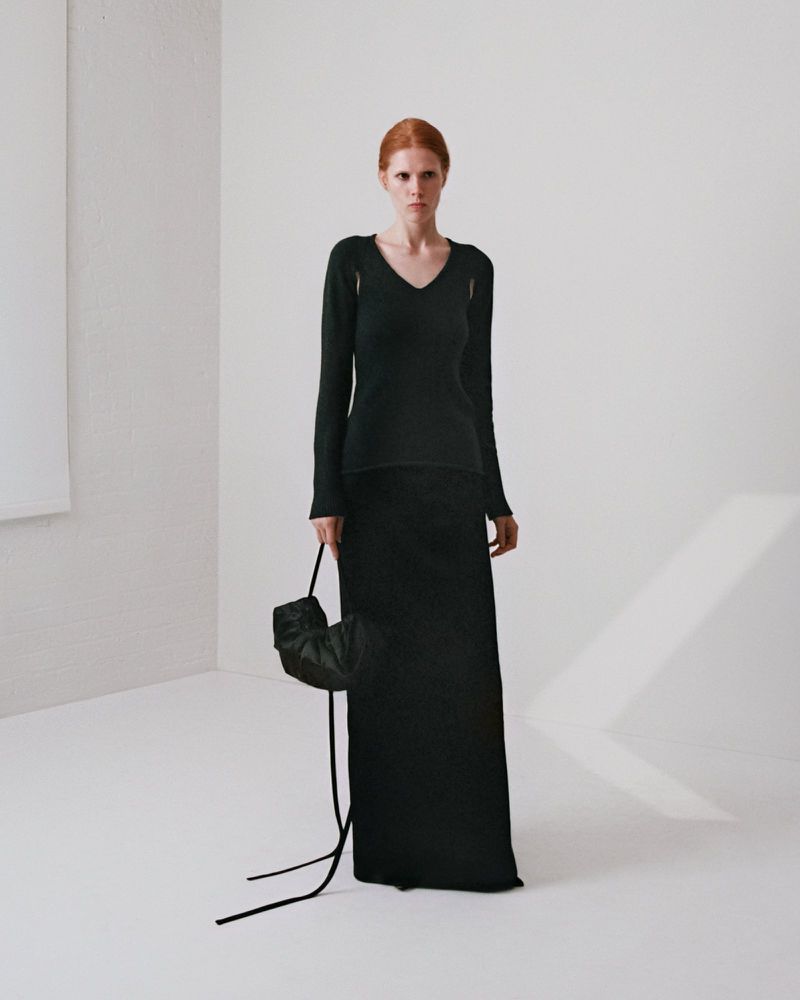 Model with red hair wearing a long black dress and long-sleeve top holding a black handbag in a white studio