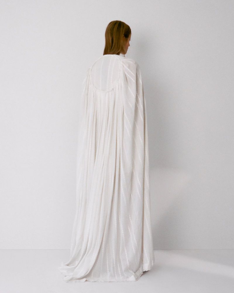 Model standing away from camera wearing a long white pleated gown against a white background