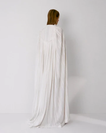 Model standing away from camera wearing a long white pleated gown against a white background