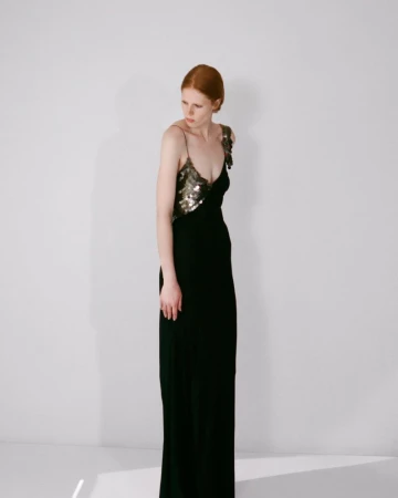 Model with red hair wearing a black evening dress with metallic embellishments against a plain white wall