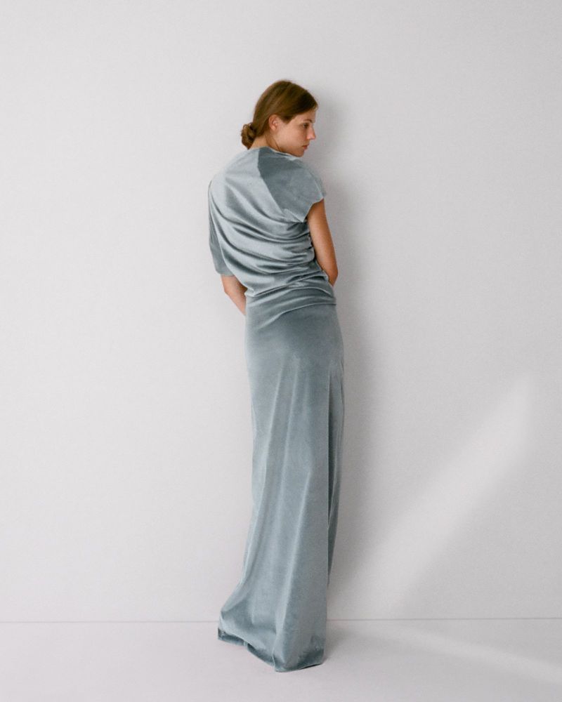 Model wearing a long light blue velvet dress standing against a plain white studio background.