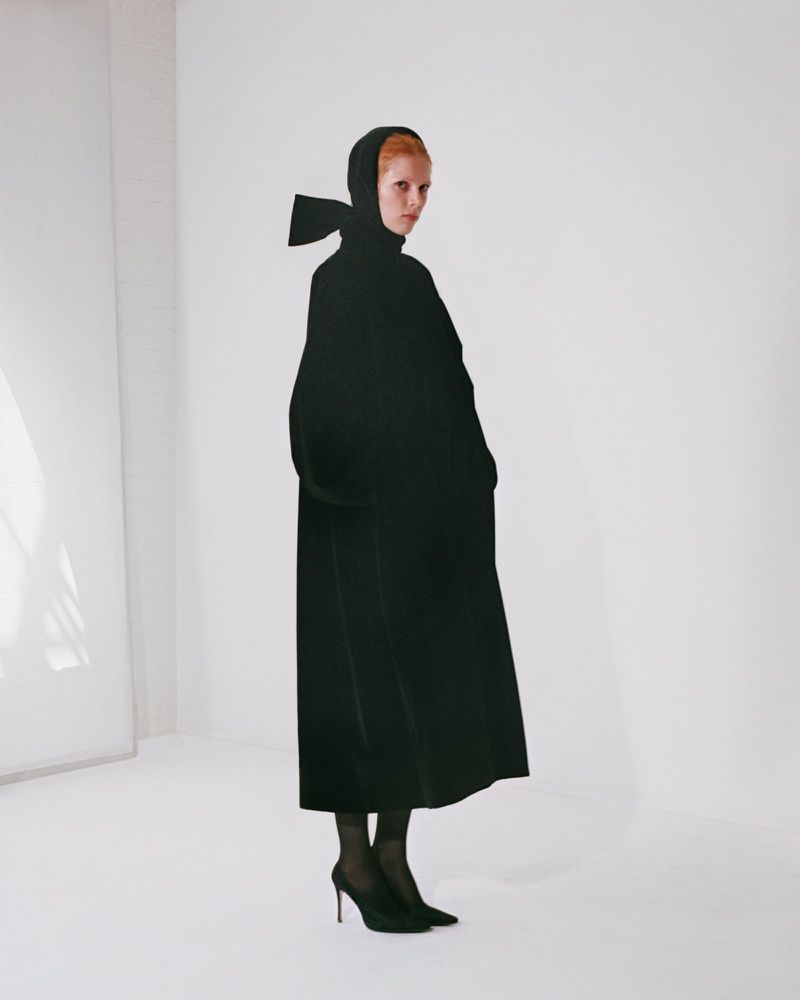 Model wearing a long black coat with a matching head covering and bow against a plain white studio backdrop