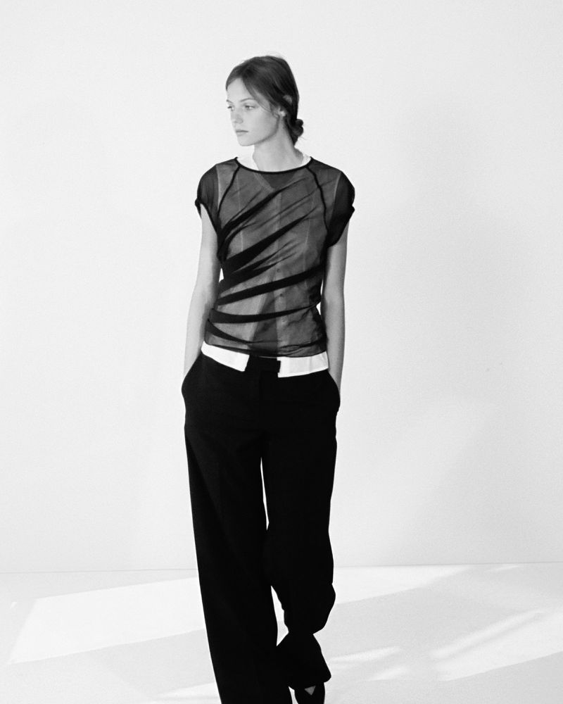 Model standing against a plain background wearing a sheer black top and dark trousers for Colleen Allen lookbook.