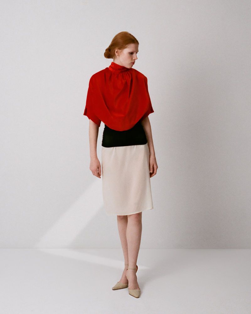 Model with red hair wearing a red draped top, black band, and cream skirt against a neutral studio backdrop.