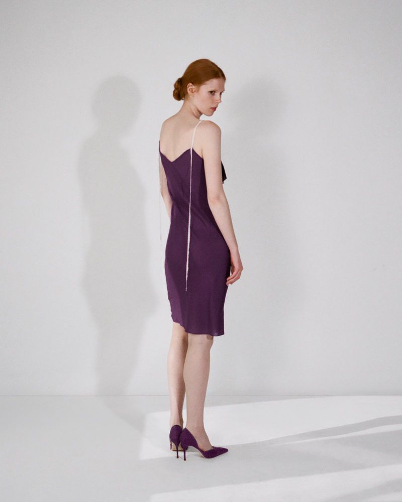 Model wearing a purple slip dress and purple heels posing against a plain white studio wall