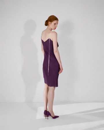 Model wearing a purple slip dress and purple heels posing against a plain white studio wall