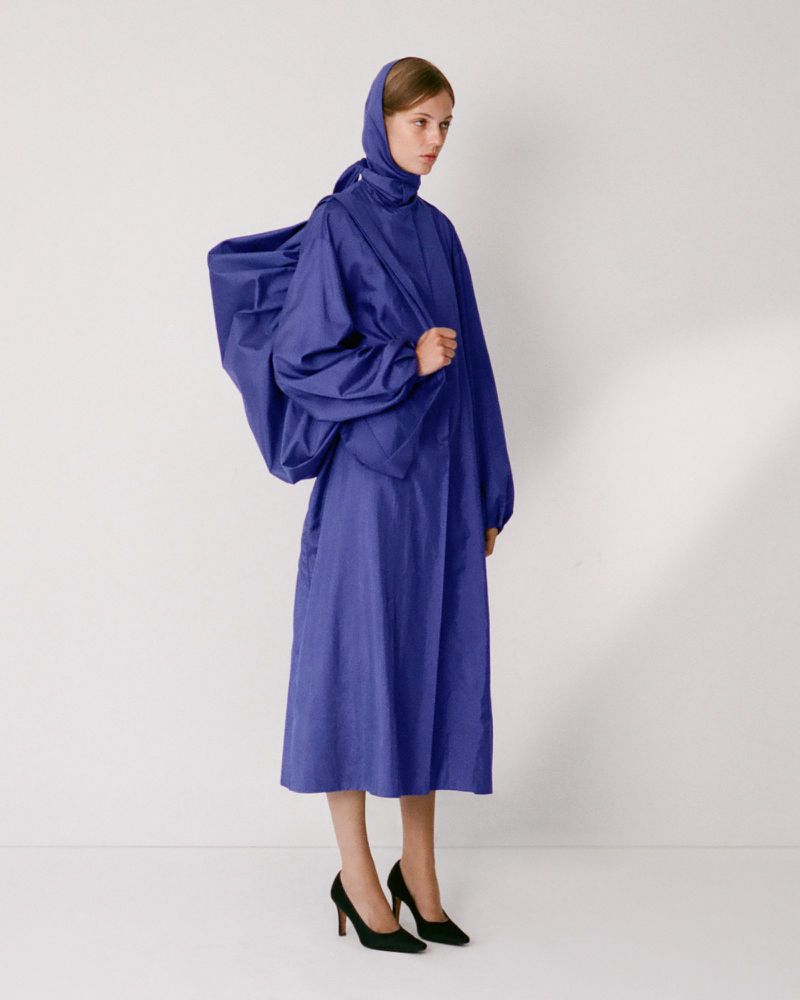 Model in royal blue hooded coat with voluminous draped back standing against a plain white background