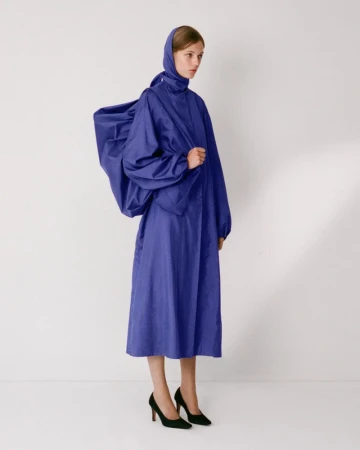 Model in royal blue hooded coat with voluminous draped back standing against a plain white background