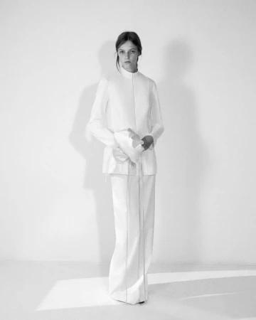 Model wearing a white structured jacket and matching trousers standing against a seamless white studio wall.
