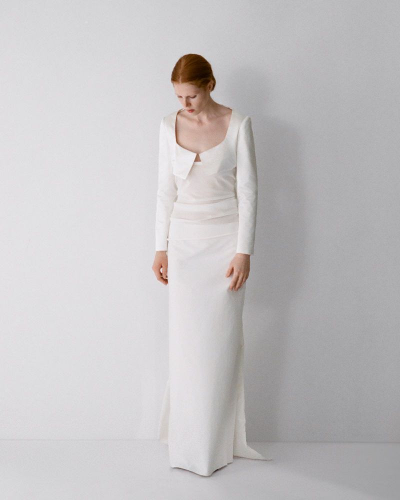 Model with red hair wearing a long white dress standing against a seamless white studio background