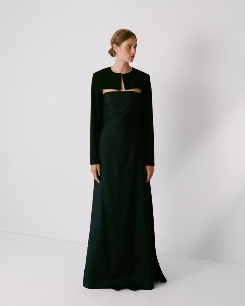 Model standing against a white backdrop wearing a dark long-sleeve bolero jacket over a matching strapless dress.