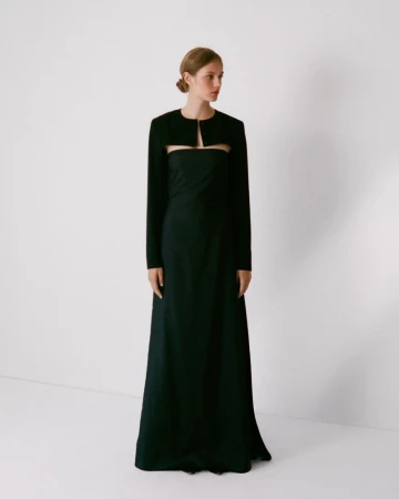 Model standing against a white backdrop wearing a dark long-sleeve bolero jacket over a matching strapless dress.