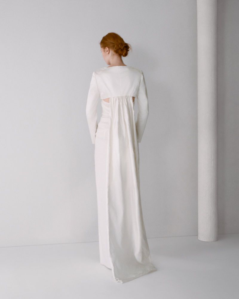 Model seen from behind wearing a white long-sleeve gown with a flowing panel against a plain studio background.