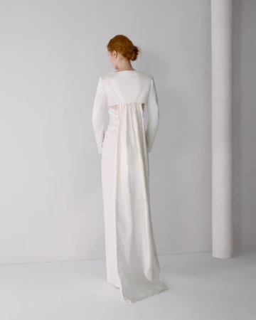 Model seen from behind wearing a white long-sleeve gown with a flowing panel against a plain studio background.