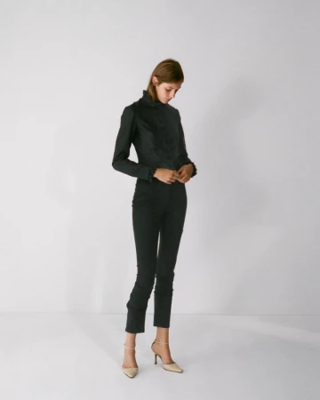 Model standing against a plain white studio background wearing a dark high-necked top and slim trousers with heeled shoes.