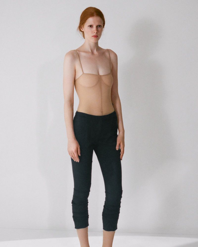 Model with red hair wearing a sheer nude top and dark pants against a white background