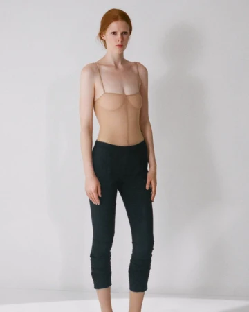 Model with red hair wearing a sheer nude top and dark pants against a white background