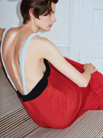 Model with short brown hair wearing a red dress with blue straps and black bodice seated indoors