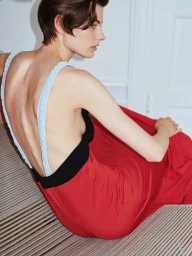 Model with short brown hair wearing a red dress with blue straps and black bodice seated indoors