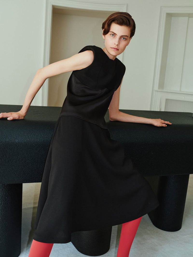 Model in black sleeveless top and midi skirt with bright red tights leaning on a dark table in an interior space