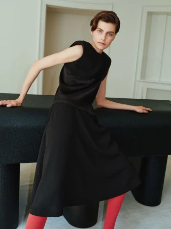 Model in black sleeveless top and midi skirt with bright red tights leaning on a dark table in an interior space