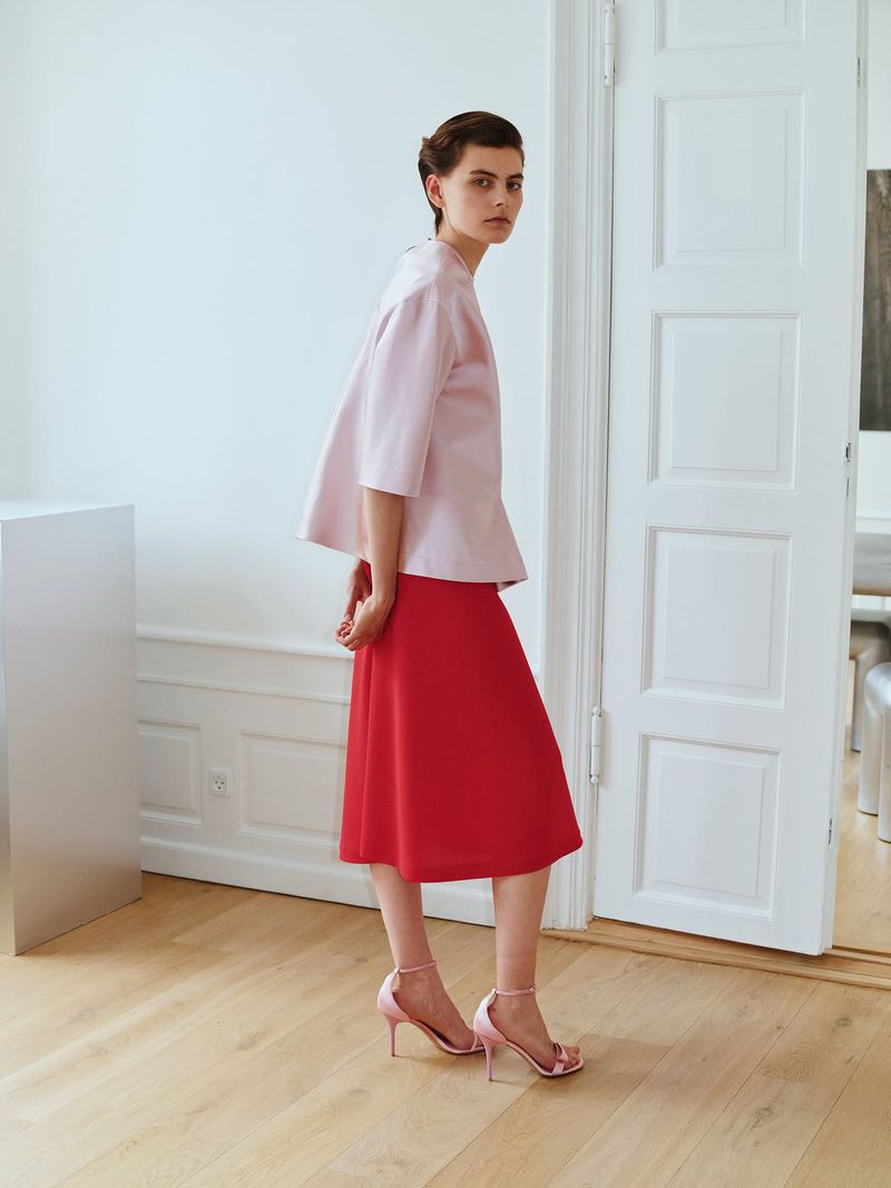 Model wearing a pink blouse and red skirt standing in a light-filled interior with white panelled doors.