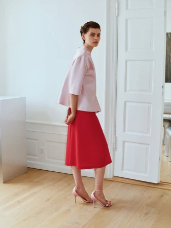 Model wearing a pink blouse and red skirt standing in a light-filled interior with white panelled doors.