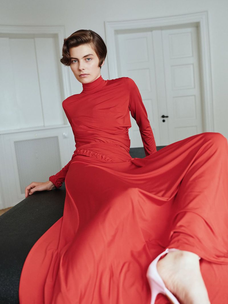Model with short dark hair wearing a vibrant red long sleeve top and flowing red skirt in a bright white interior setting