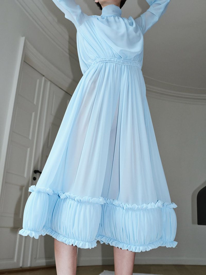 Model in a light blue pleated midi dress with high neck and ruffled hem posing indoors with arms raised