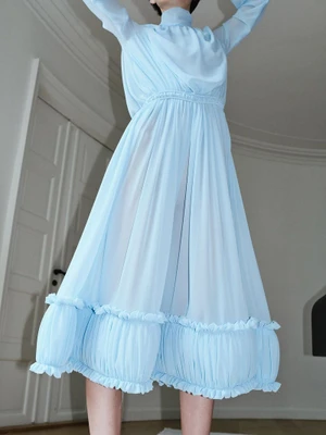Model in a light blue pleated midi dress with high neck and ruffled hem posing indoors with arms raised