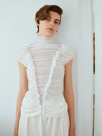 Model wearing a white pleated sheer high-neck top standing against a pale wall for Forza Collective