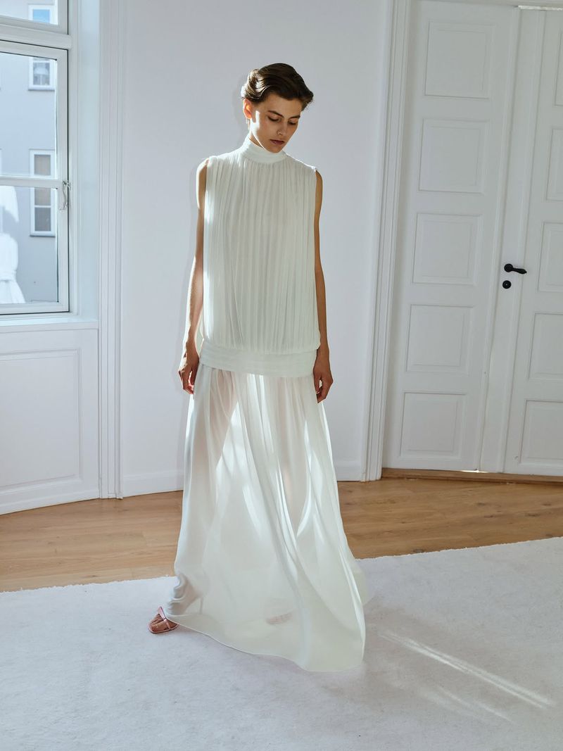 Model wearing a white sleeveless pleated top and sheer long skirt standing inside a bright room