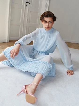 Model seated on the floor wearing a light blue chiffon dress and pink high heel sandals in a room with white doors.