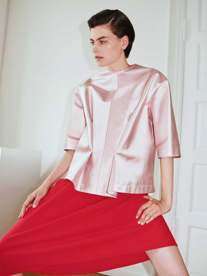 Model with short dark hair wearing a pink satin top and bright red skirt in a bright indoor setting