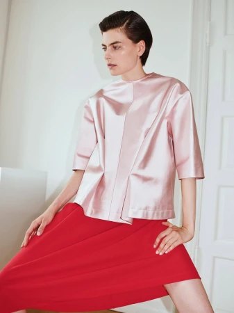Model with short dark hair wearing a pink satin top and bright red skirt in a bright indoor setting