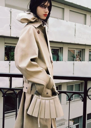 Model in a beige trench coat holding a pleated handbag on a balcony for Lacoste