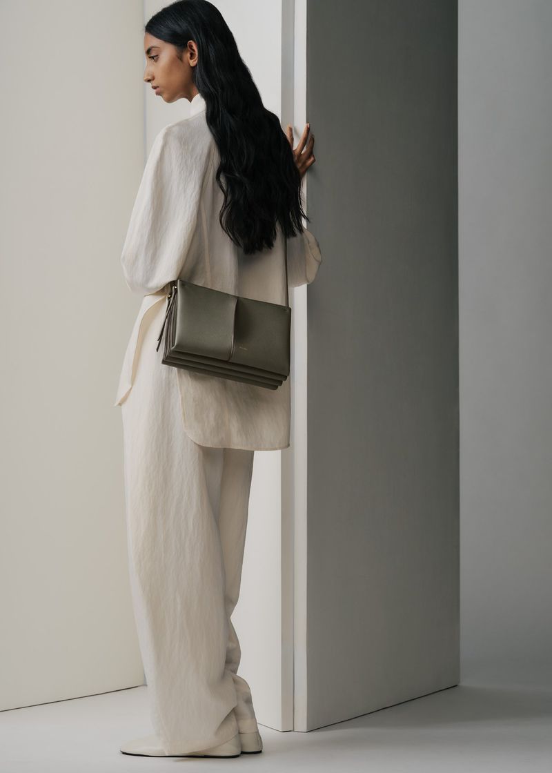 Model with long dark hair wearing white clothing and carrying an olive green shoulder bag against a neutral background