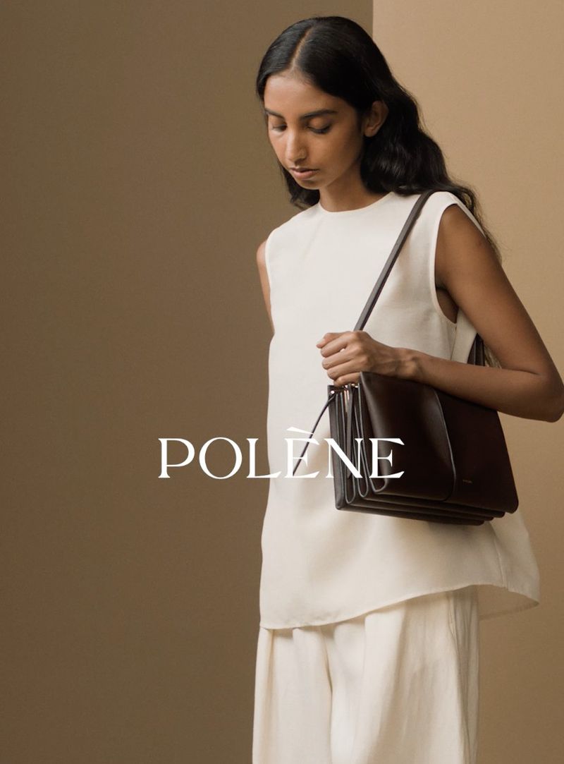 Model in white sleeveless top and trousers holding a dark brown leather shoulder bag against a neutral background
