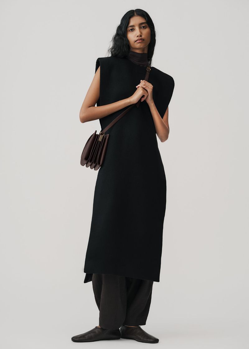 Model wearing a black sleeveless dress and dark trousers holding a brown leather crossbody bag against a plain grey backdrop.