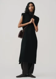 Model wearing a black sleeveless dress and dark trousers holding a brown leather crossbody bag against a plain grey backdrop.