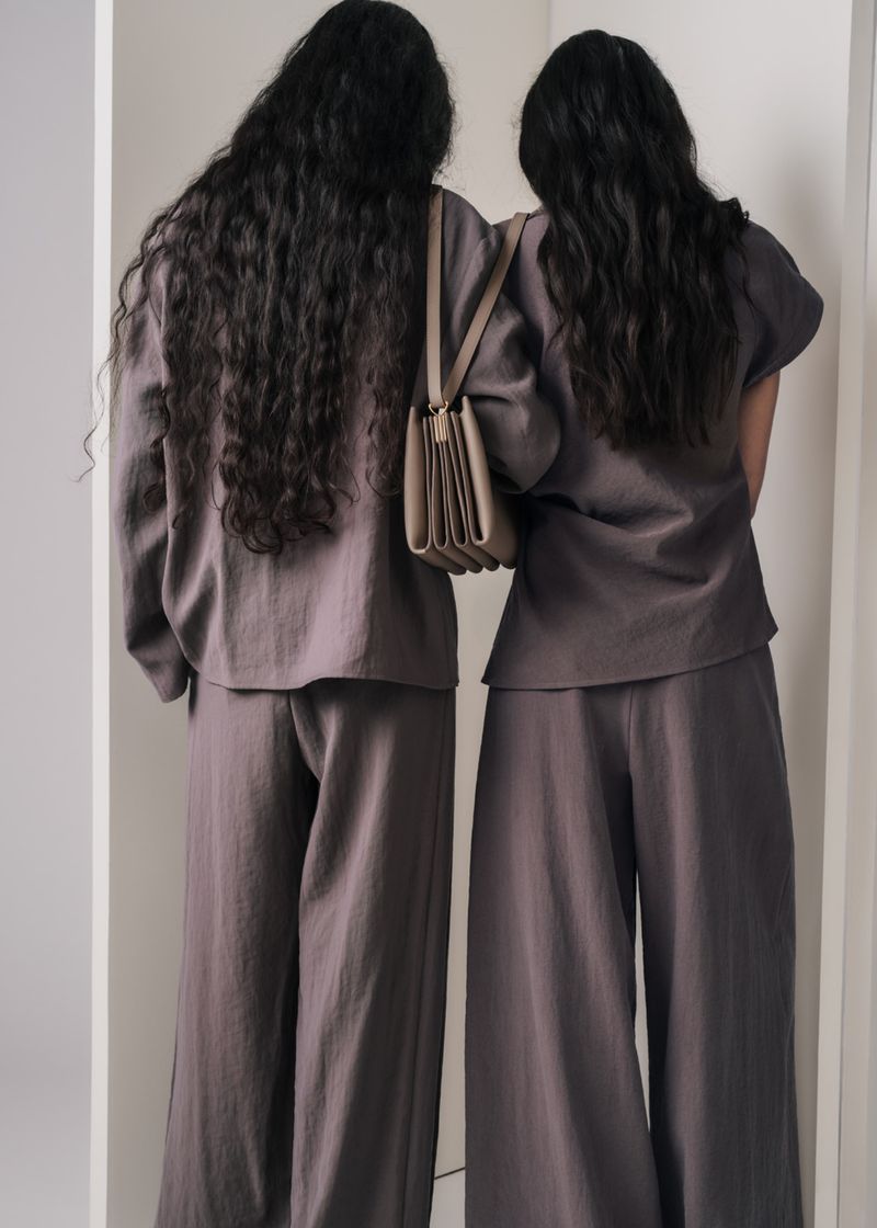 Two women with long dark hair wearing matching grey outfits carrying a tan shoulder bag against a neutral studio backdrop
