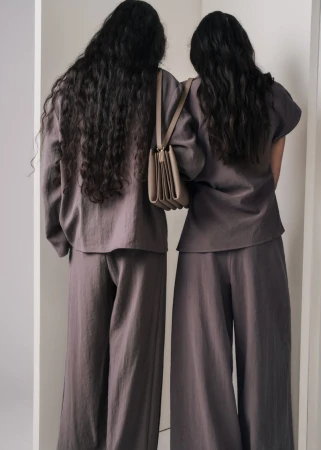 Two women with long dark hair wearing matching grey outfits carrying a tan shoulder bag against a neutral studio backdrop