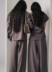 Two women with long dark hair wearing matching grey outfits carrying a tan shoulder bag against a neutral studio backdrop