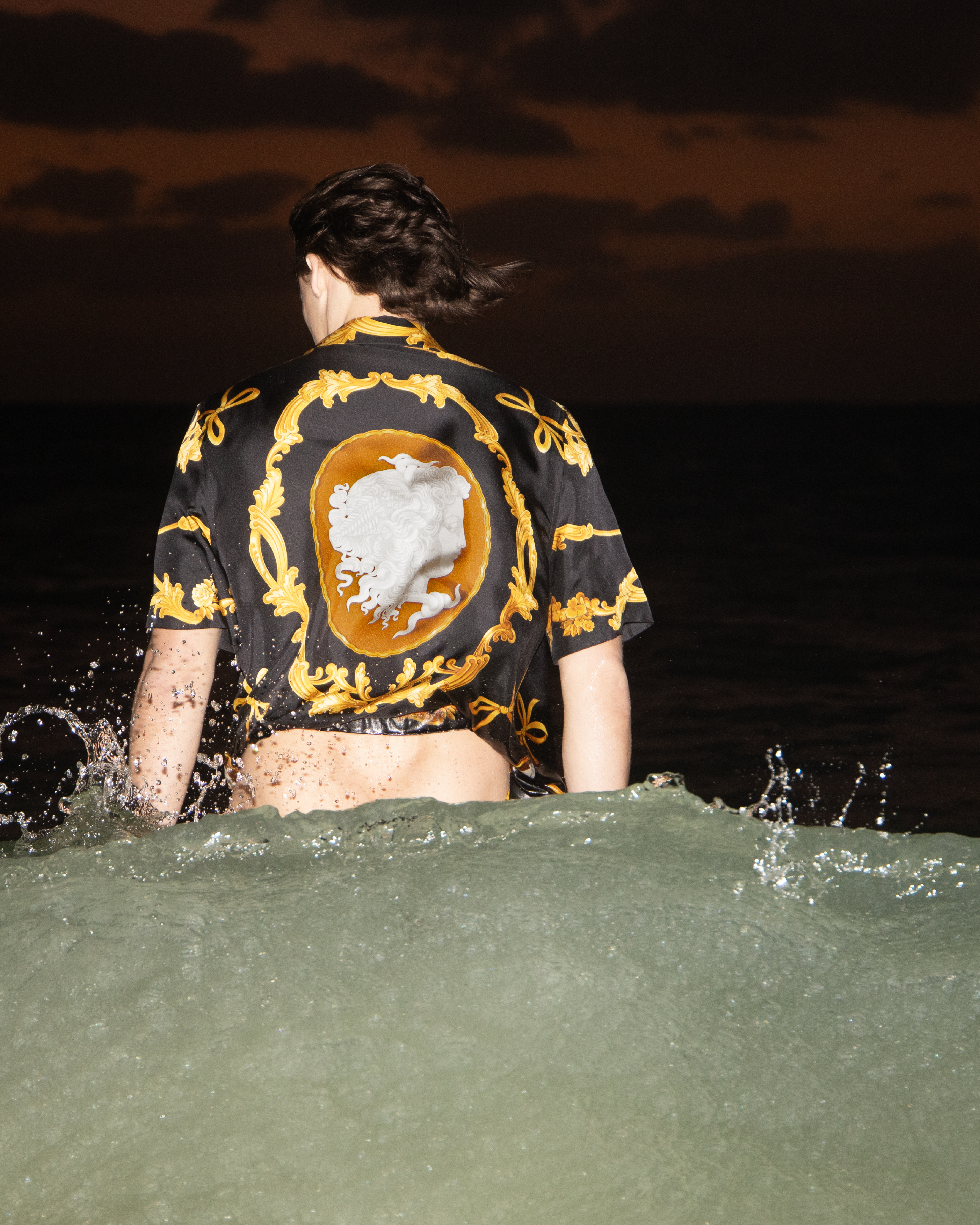 Person in water at dusk, wearing a patterned black and gold shirt. The ocean wave is visible in the foreground.