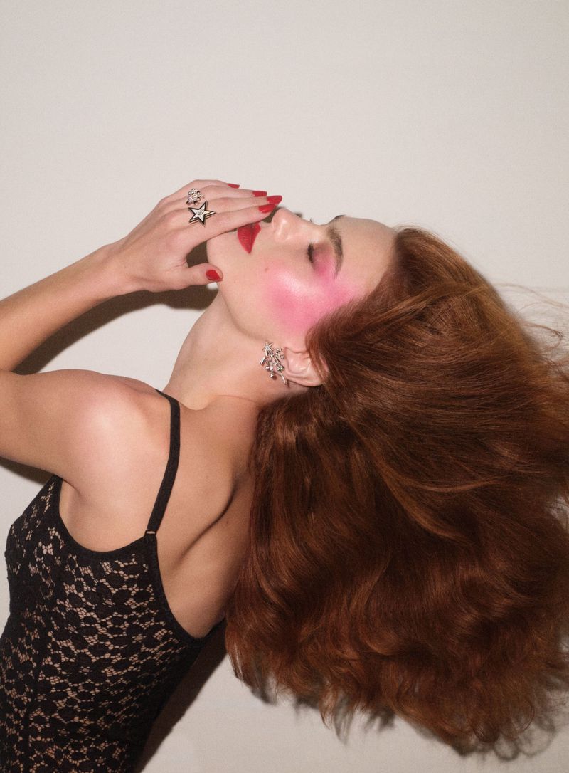 Adult female model with red hair in a black lace top poses with red lipstick and blush against a white background in a beauty fashion shot.