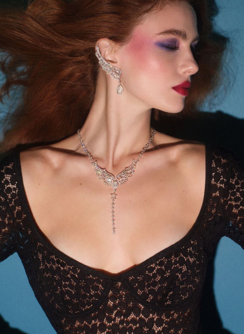 Portrait of an adult woman with red hair, wearing a black lace top and diamond jewelry, with dramatic eye and lip makeup against a blue background.
