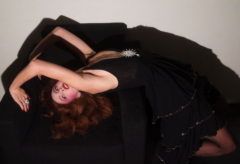 A woman with red hair poses in a black dress on a couch. She is angled to the right with her head upside down and her arms up.