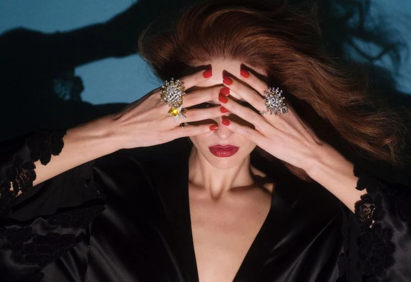 Woman with auburn hair wearing black robe and oversized rings, hands covering her face, striking red lipstick, against a dark blue background.